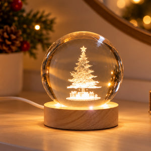 A glowing Christmas tree snow globe lamp with a warm light inside a clear sphere, set on a round wooden base on a tabletop. A blurred holiday plant appears in the background.