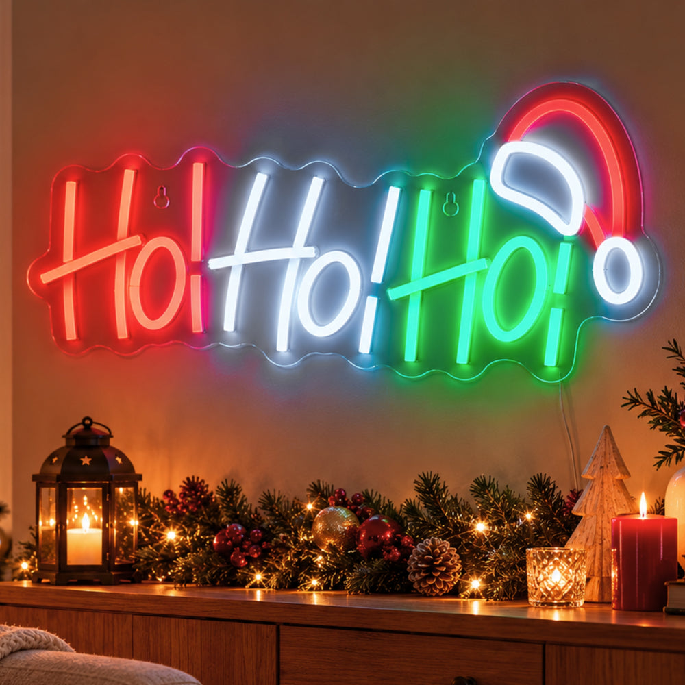 A glowing neon-style wall sign spelling "Ho Ho Ho" in red, white, and green, mounted above a holiday-decorated mantel with candles, pinecones, ornaments, and a small Christmas tree in a warmly lit room.