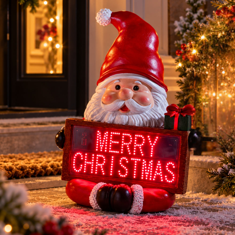 A festive Santa Claus garden statue holding a glowing red LED sign that reads “MERRY CHRISTMAS,” placed outdoors in front of a decorated holiday home with warm lights and a Christmas tree.