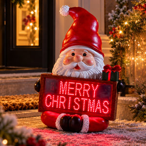 A festive Santa Claus garden statue holding a glowing red LED sign that reads “MERRY CHRISTMAS,” placed outdoors in front of a decorated holiday home with warm lights and a Christmas tree.