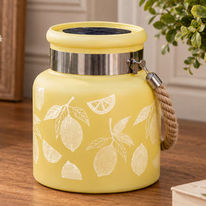 A yellow glass jar with a lemon pattern, featuring a metal band, a rope handle, set on a wooden table.