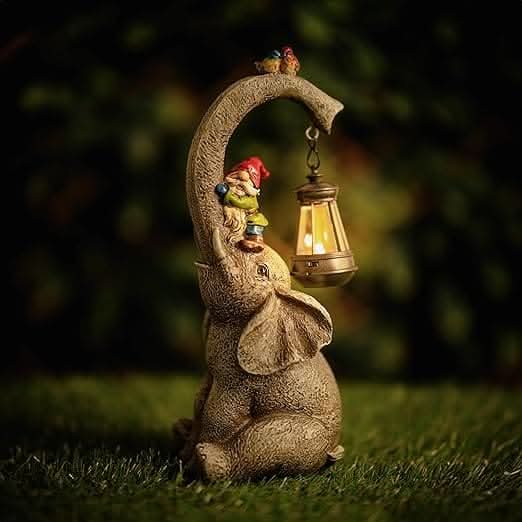 A solar garden ornament featuring a stone elephant sitting on grass with a small figurine of a person on its back holding a lantern, at dusk.