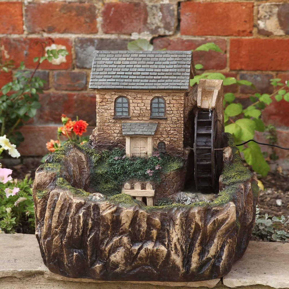 A miniature stone mill garden ornament with a tiny house, water wheel, and faux rock basin, displayed outdoors against a brick wall and garden plants.