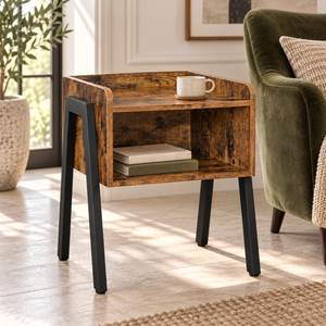 A rustic wooden end table with black metal legs, styled in a bright living room next to a green armchair with a coffee cup on top.