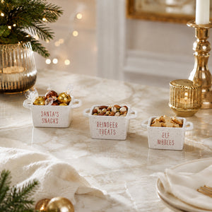 Three small white ceramic snack bowls with handles labeled 'Santa's Snacks', 'Reindeer Treats', and 'Elf Nibbles' filled with assorted chocolates and nuts, styled on a marble tabletop with holiday decor.