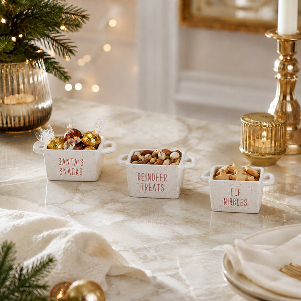 Three small white ceramic snack bowls with handles labeled 'Santa's Snacks', 'Reindeer Treats', and 'Elf Nibbles' filled with assorted chocolates and nuts, styled on a marble tabletop with holiday decor.