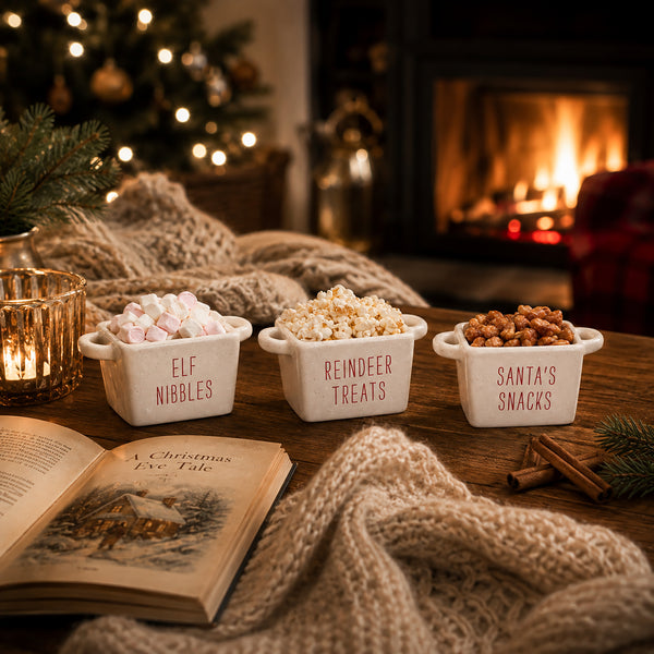 Three white ceramic snack bowls labeled 'ELF NIBBLES', 'REINDEER TREATS', and 'SANTA'S SNACKS' filled with marshmallows, popcorn, and peanuts, arranged on a wooden table in a cozy holiday setting with a knitted blanket, fireplace, and open book.
