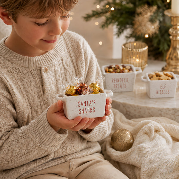 A child in a cream cable-knit sweater holds a small white ceramic bowl labeled 'SANTA'S SNACKS' filled with wrapped chocolates. In the background, a holiday table displays other labeled snack bowls, including 'REINDEER TREATS' and 'ELF NIBBLES', with a softly lit Christmas tree and candles.