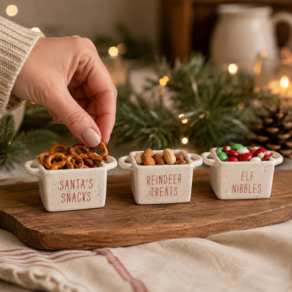 Three small white ceramic snack bowls labeled 'Santa's Snacks', 'Reindeer Treats', and 'Elf Nibbles' on a wooden board, with a hand reaching for a pretzel from the left bowl in a cozy holiday setting.