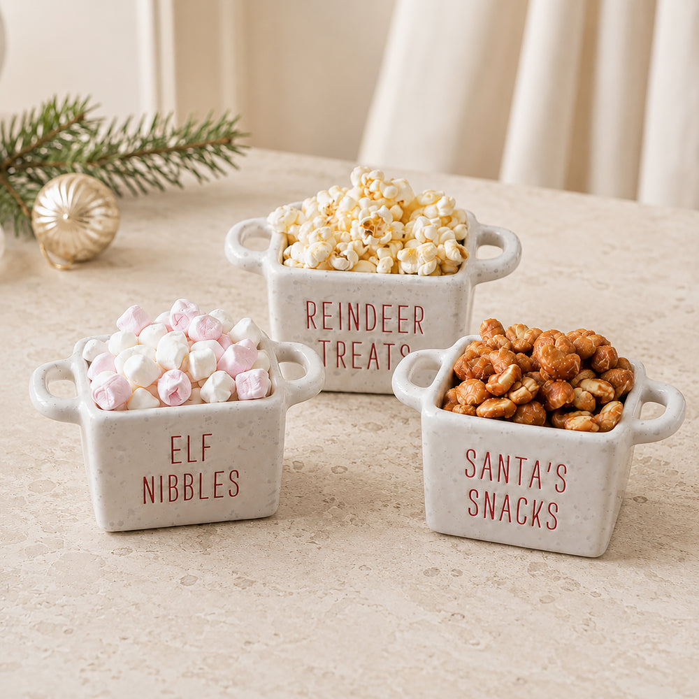 Three small white ceramic snack bowls with handles sit on a beige tabletop. The bowls are labeled 'ELF NIBBLES,' 'REINDEER TREATS,' and 'SANTA'S SNACKS' in red lettering and are filled with marshmallows, popcorn, and caramel-coated nuts.