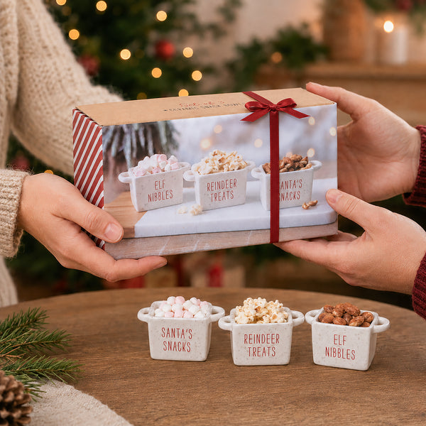 A gift box tied with a red ribbon is held by two hands over a wooden table. The box features a printed photo of three small white snack bowls labeled 'Elf Nibbles', 'Reindeer Treats', and 'Santa's Snacks'.