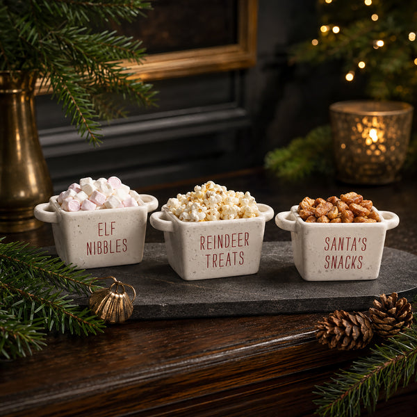 Three small white ceramic snack bowls with handles labeled 'ELF NIBBLES', 'REINDEER TREATS', and 'Santa's Snacks' filled with marshmallows, popcorn, and nuts, arranged on a dark stone tray in a cozy holiday setting.