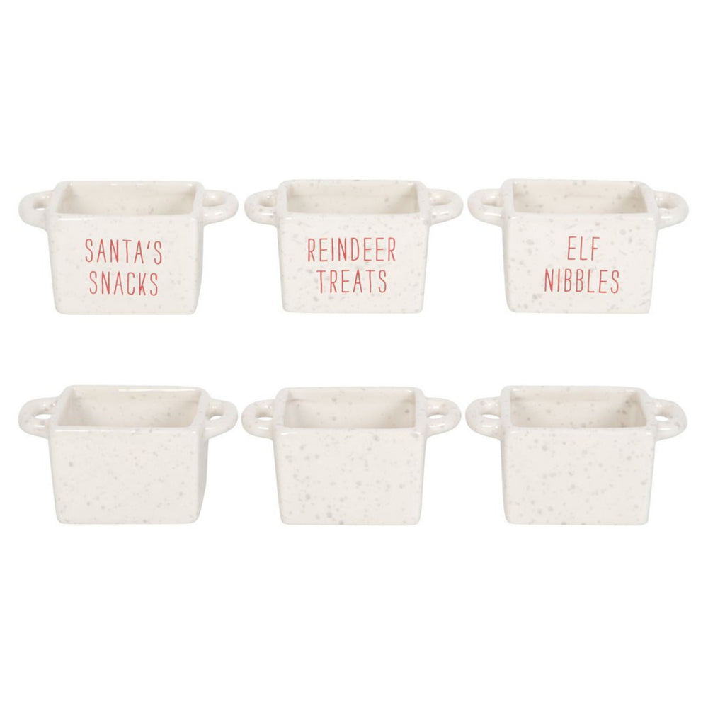 Three small white ceramic snack bowls with handles sit on a beige tabletop. The bowls are labeled 'ELF NIBBLES,' 'REINDEER TREATS,' and 'SANTA'S SNACKS' in red lettering and are filled with marshmallows, popcorn, and caramel-coated nuts.