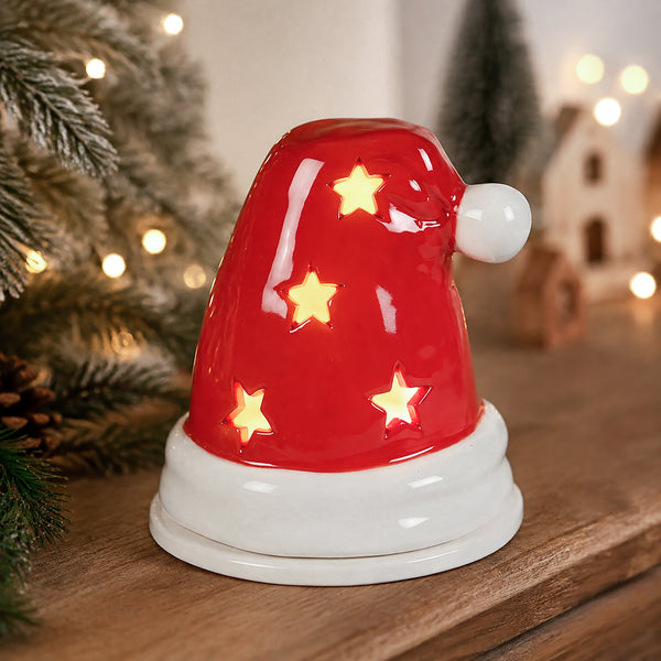 A red Santa hat-shaped ceramic candle holder with glowing star cutouts, displayed on a wooden surface with a blurred Christmas background.