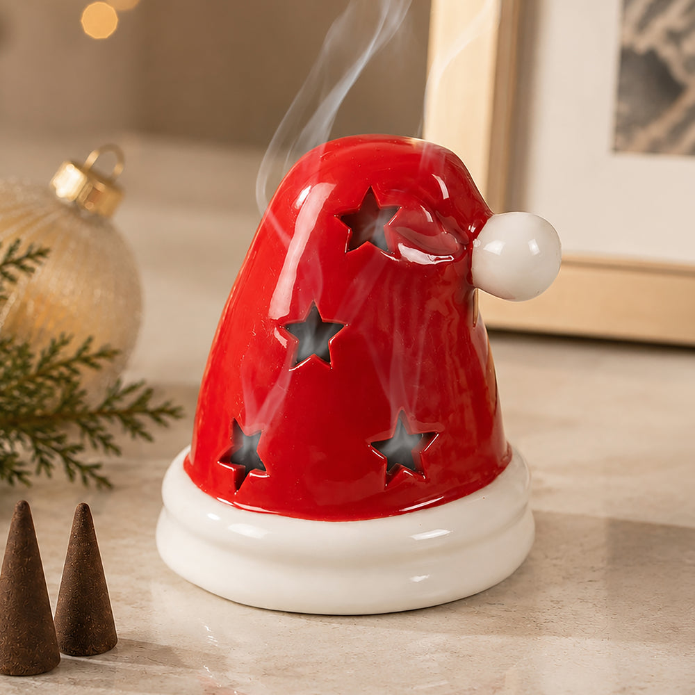 A red Santa hat-shaped ceramic diffuser with star cutouts and a white base, shown with smoke rising from the top on a tabletop.