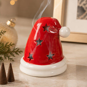 A red Santa hat-shaped ceramic diffuser with star cutouts and a white base, shown with smoke rising from the top on a tabletop.