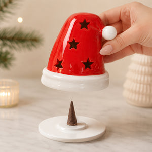 A hand holds a red Santa hat-shaped incense holder above a white base with a small brown incense cone, set on a light marble surface with soft holiday decor in the background.