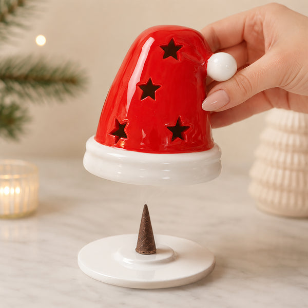 A hand holds a red Santa hat-shaped incense holder above a white base with a small brown incense cone, set on a light marble surface with soft holiday decor in the background.