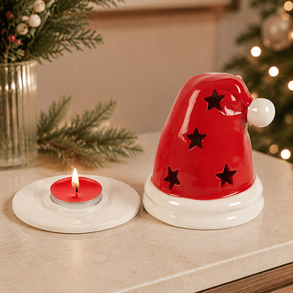 A red Santa hat-shaped candle holder with black star cutouts and a white pom-pom, shown beside a small lit tealight candle on a white saucer on a tabletop. Christmas greenery and a blurred lit tree appear in the background.