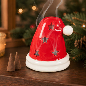 A red Santa hat-shaped diffuser or incense holder with star cutouts, shown on a wooden surface with smoke rising and holiday greenery in the background.