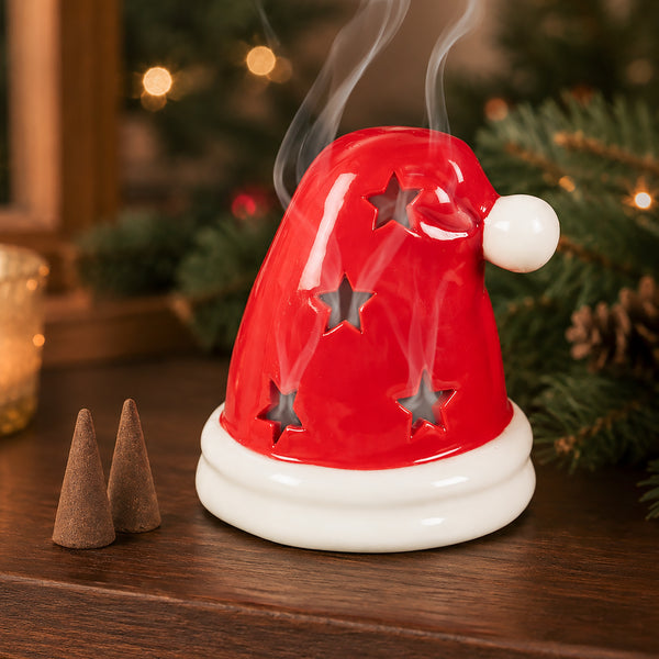 A red Santa hat-shaped diffuser or incense holder with star cutouts, shown on a wooden surface with smoke rising and holiday greenery in the background.