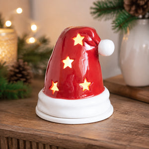 A red Santa hat-shaped tea light candle with glowing star cutouts and a white pom-pom, displayed on a wooden surface with festive holiday decorations in the background.
