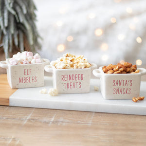Three small bowls labeled 'Elf Nibbles', 'Reindeer Treats', and 'Santa's Snacks' on a table with a festive background.