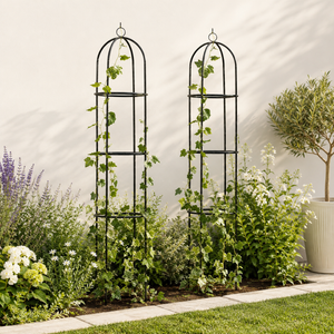 Two black metal garden trellises with arched tops, shown in a landscaped garden setting with green ivy climbing them, surrounded by various green plants and flowers against a light beige wall.