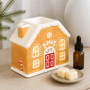 A gingerbread house-shaped ceramic candle holder with a red door, white snow-dusted roof, and candy cane detail, styled on a wooden surface with a small dish of wax cubes and an amber dropper bottle nearby.