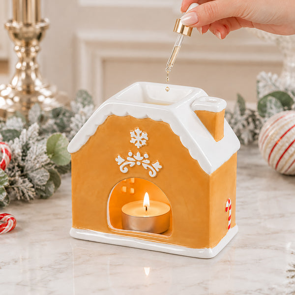 A gingerbread house-shaped candle holder with a lit tealight candle inside, shown on a marble surface with holiday decorations. A hand is holding a glass dropper above the top compartment, suggesting fragrance oil addition.