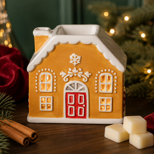 A gingerbread house-shaped ceramic candle holder or wax warmer with a warm yellow-orange body, white snow-dusted roof and trim, a red door, and decorative window details, styled on a wooden surface with holiday decor and lit Christmas trees in the background.