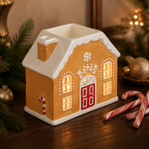A gingerbread house-shaped ceramic container with a warm yellow-orange body, white roof and trim, a red door, and glowing yellow window lights, styled on a wooden surface with holiday decorations including candy canes, evergreen branches, and ornaments.