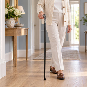 An elderly woman walking indoors holding a black cane with a decorative silver handle. She is wearing a cream cardigan, white top and trousers, and brown loafers in a bright, modern hallway with light wood floors.