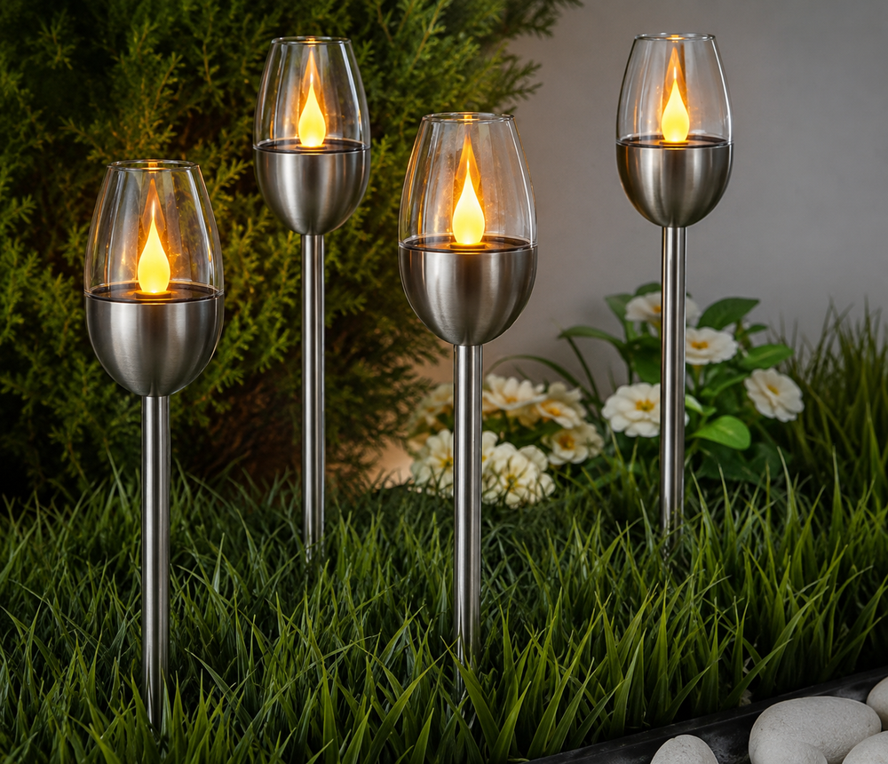 Four stainless steel solar flame torches with clear glass cups are arranged in green grass outdoors, with artificial-looking flames lit inside each holder.