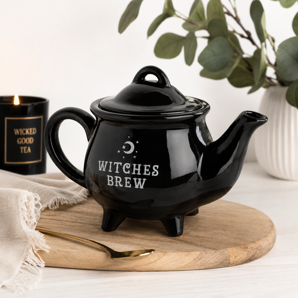 Glossy black Witches Brew teapot with crescent moon design, displayed on a wooden tray with gold spoon and candle.
