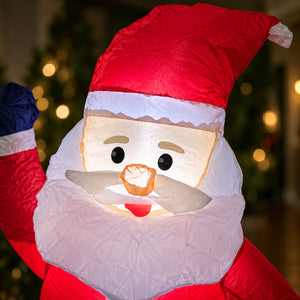 A close-up of a Santa Claus inflatable or decorative figure with a red hat, white beard, and glowing face, photographed against a blurred holiday background with lights.