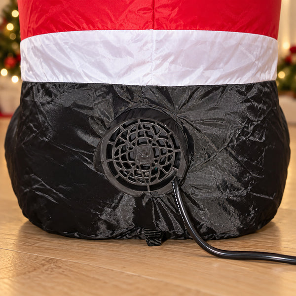 A close-up of a black inflatable pump attachment connected to an inflatable Christmas decoration, with the decoration showing a red and white top section. The pump cord is visible on a wooden floor with blurred Christmas lights in the background.