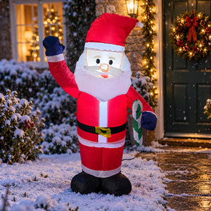 A large inflatable Santa Claus decoration stands in the snow outside a decorated house. Santa is wearing a red suit with white trim, a black belt, blue gloves, and a red hat, and is holding a green gift box.