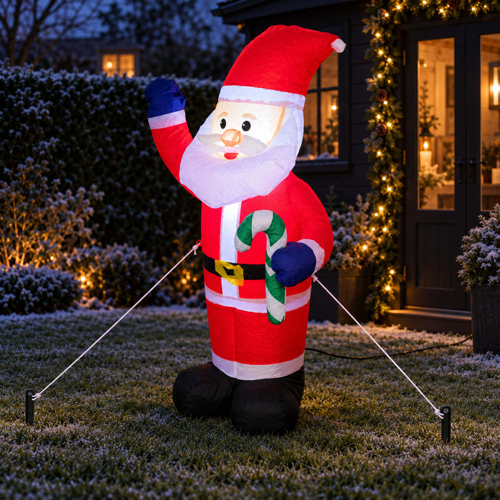A large inflatable Santa Claus yard decoration stands on a lawn in front of a house at dusk. The inflatable Santa wears a red suit with white trim, blue gloves, and holds a green-and-white candy cane.