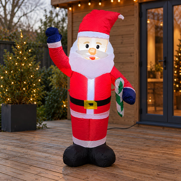 An inflatable outdoor Santa Claus decoration stands on a wooden deck in front of a house. The Santa figure is red with white trim, a black belt, blue gloves, and holds a green and white candy cane.