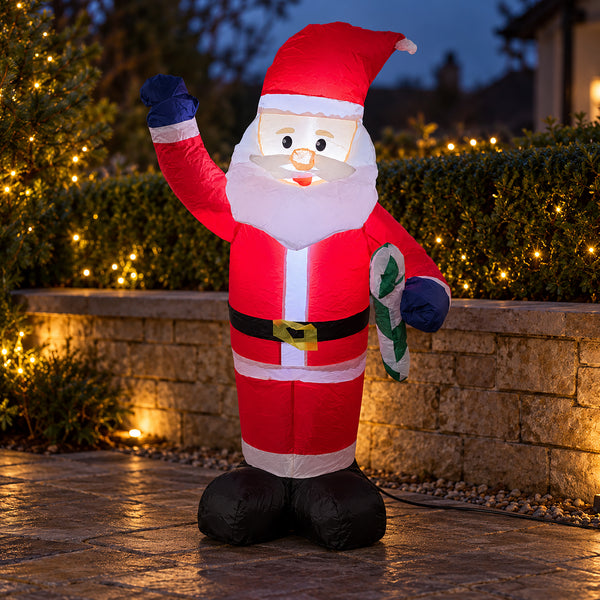 A large inflatable Santa Claus outdoor decoration stands on a stone walkway at dusk, with lit hedges and a stone wall behind it. The Santa figure is red with white trim, a black belt, dark blue gloves, and holds a green-and-white candy cane.