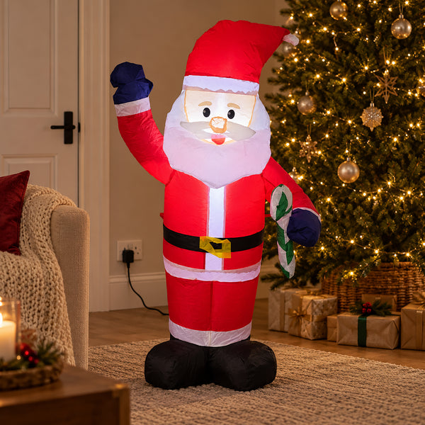 An inflatable Santa Claus decoration stands in a living room beside a lit Christmas tree and wrapped gifts. The Santa figure is red with a white beard and cuffs, black boots and belt, and blue gloves, holding a green gift bag.