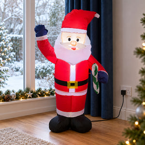 An inflatable Santa Claus figure stands on a wooden floor in a decorated room, with a snowy window, blue curtains, and a Christmas tree visible in the background.
