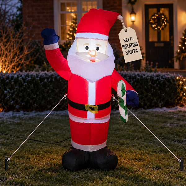 An inflatable Santa Claus yard decoration stands on a lawn at dusk, holding a sign that reads 'SELF-INFLATING SANTA.' The figure is dressed in a red suit with white trim, black belt and boots, and blue gloves, with a white beard.