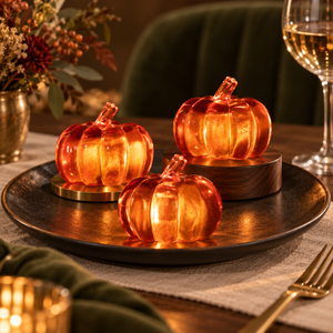 Three small translucent orange glass pumpkins with gold-toned bases, arranged on a dark round platter in a warm dining setting with a wine glass, flowers, and green table textiles in the background.
