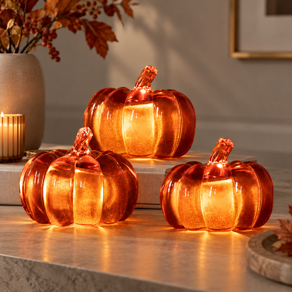 Three amber-orange glass pumpkins with textured interiors arranged on a tabletop in a warmly lit indoor setting.