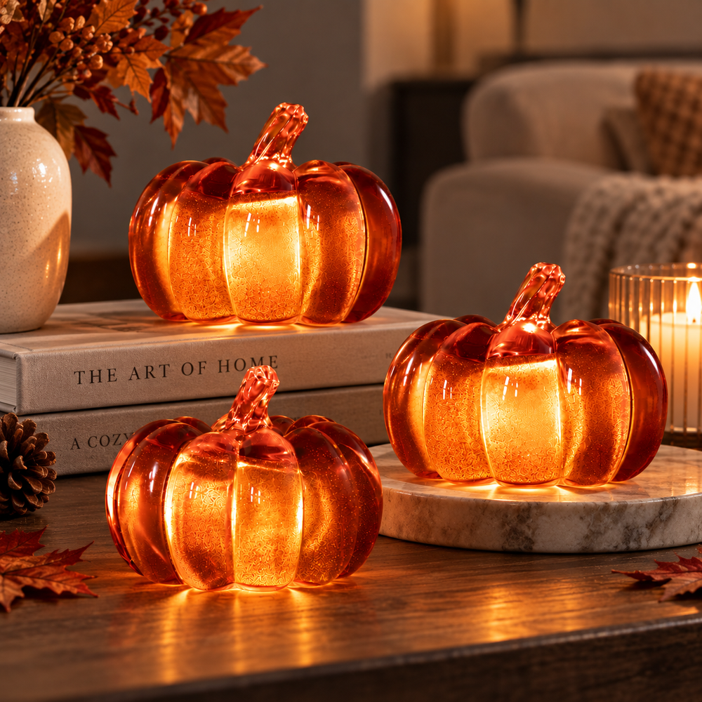 Three decorative glass pumpkins with warm orange and amber lighting, styled on a wooden table in a cozy living room setting with books, a candle, and fall leaves.
