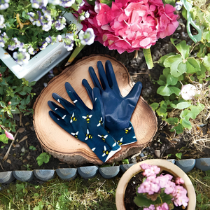 A pair of navy blue gardening gloves with yellow and white bumblebee prints, displayed on a rustic wooden slice in a garden setting with flowers.