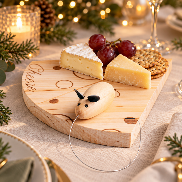 A wooden cheese board with engraved cheese shapes and a small wooden cheese puller, styled with brie, cheddar, and crackers on a table with candles, pine branches, and warm string lights in the background.