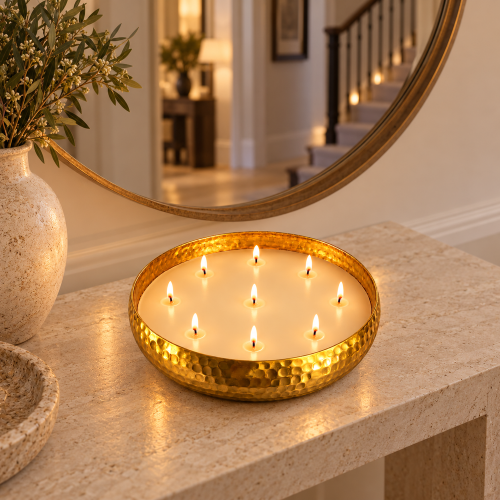 A hammered gold metal candle holder containing multiple lit candles, styled on a beige stone console table in a warm, elegant interior setting with a large round mirror and a vase in the background.